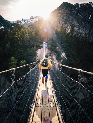 Person with a backpack crossing a suspension bridge toward the mountains with sunlight breaking through the peaks.