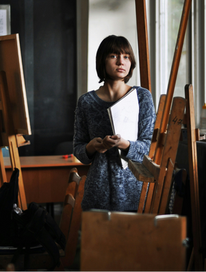 Young woman holding a sketchbook in an art studio surrounded by easels, looking thoughtful and inspired.