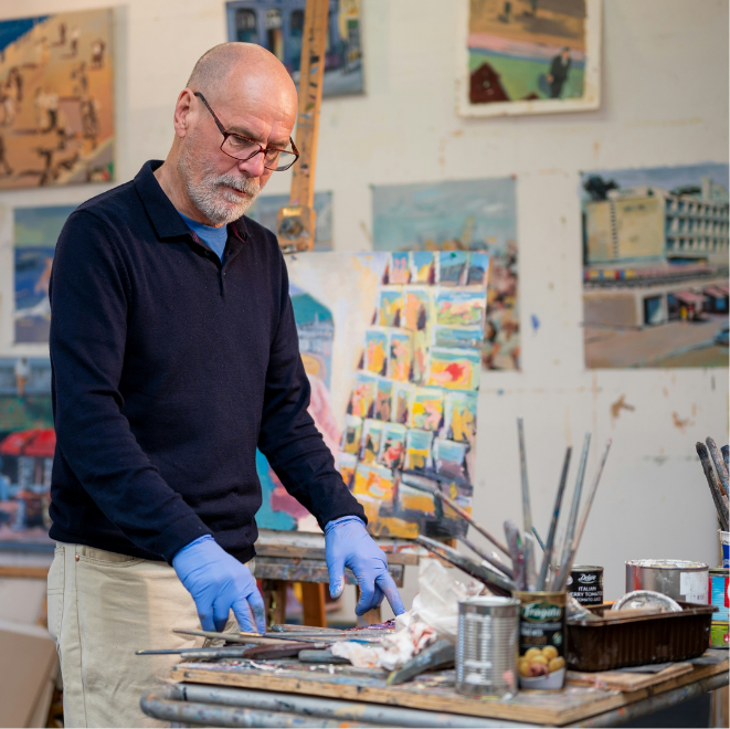 Artist in a studio wearing gloves, standing at a workbench with paints and brushes, surrounded by colourful paintings.