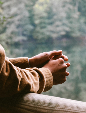 Person leaning on a wooden railing with hands clasped, gazing thoughtfully over a calm forest lake.