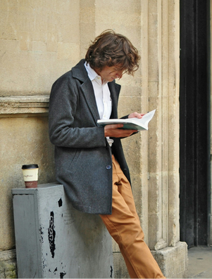 Young man in a coat leaning against a wall, reading a book intently with a takeaway coffee cup nearby.