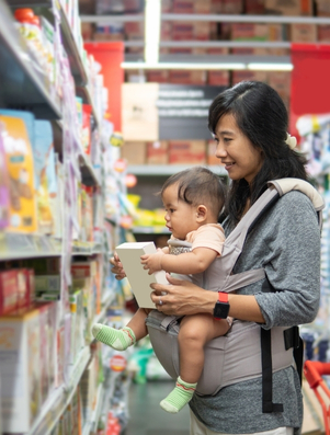 A smiling mother carrying her baby in a store aisle while looking at family products on the shelf