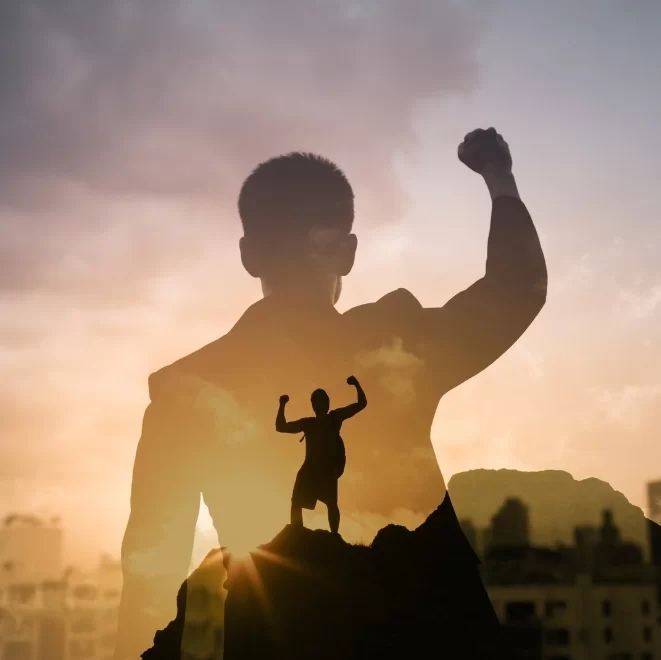 Silhouette of a man raising his fist in triumph with a double exposure of a person standing on a mountaintop in victory pose.