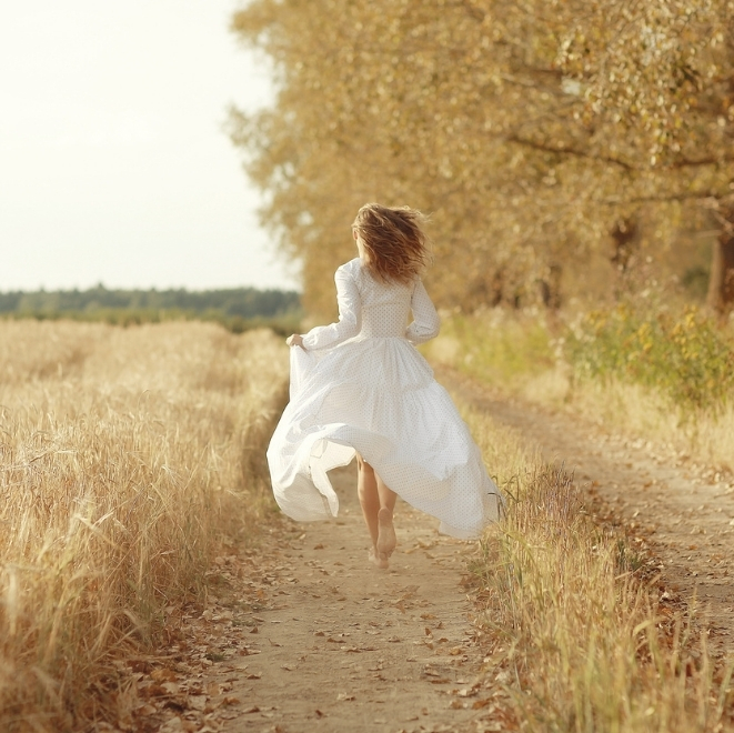Woman in a flowing white dress running barefoot along a golden field path, symbolising purity and freedom.