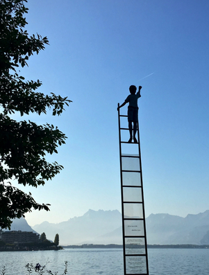 Silhouette of a person standing on top of a tall ladder reaching toward the sky, with mountains and a lake in the background.
