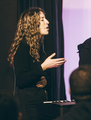 Young woman speaking passionately on stage with a microphone headset, gesturing with her hand as an audience listens.