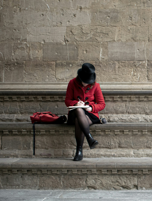 Person in a red coat and black hat sitting alone on stone steps, writing in a notebook with a red bag placed nearby.