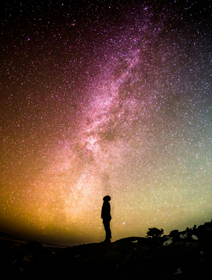 Silhouette of a person standing beneath a vast starry sky and colourful Milky Way, gazing upward in wonder.