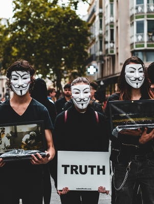 Group of people wearing Guy Fawkes masks holding signs and photos, with one sign boldly displaying the word 'TRUTH' during a street protest.
